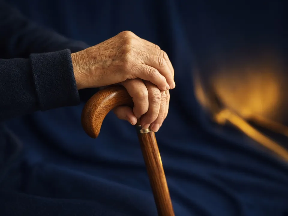 Anonymous elderly hands resting on a wooden walking cane, warm gold light